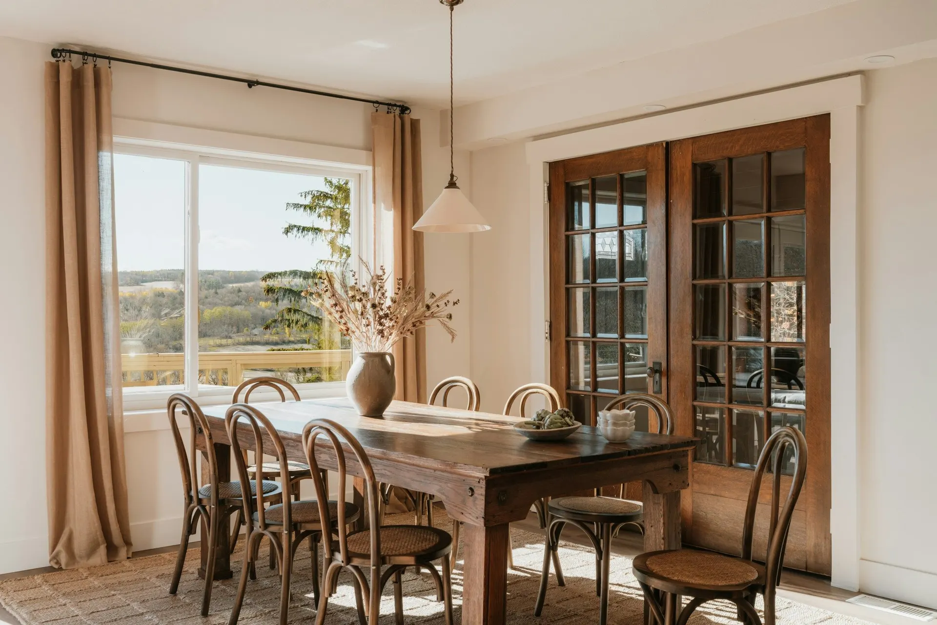 A sunlit dining room with a wood table and a window onto the countryside